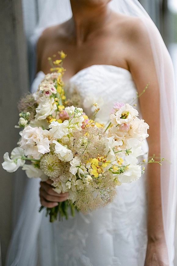 Bridal bouquet of white and yellow flowers with blush blooms and greenery held against a strapless wedding dress in soft window light