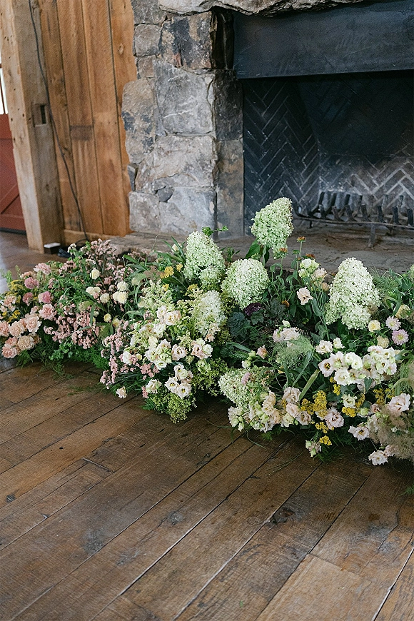 Wedding floral installation with ground floral arrangement of white hydrangeas, blush blooms, and greenery around a stone fireplace indoors