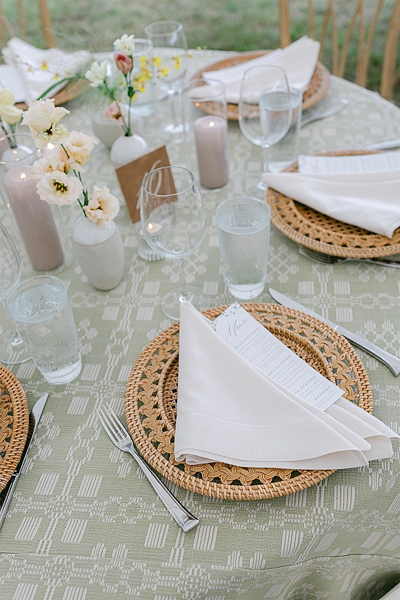 Reception tablescape with outdoor wedding tablescape details, rattan chargers, white napkins, menus, pastel bud vases and gray candles on a green patterned cloth outdoors