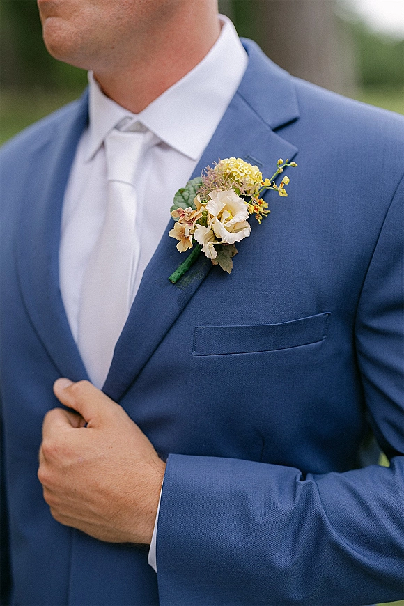 Groom boutonniere with yellow and white flowers pinned to a blue suit jacket, crisp white tie and shirt, against blurred greenery backdrop