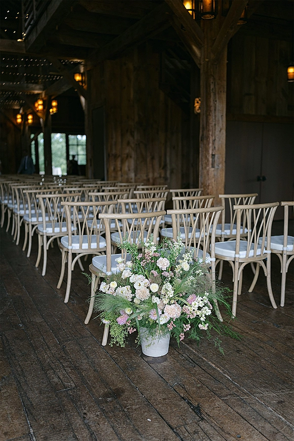 Ceremony seating with wedding ceremony chair rows of wood chairs and cushions lining a rustic barn aisle with flowers and string lights overhead