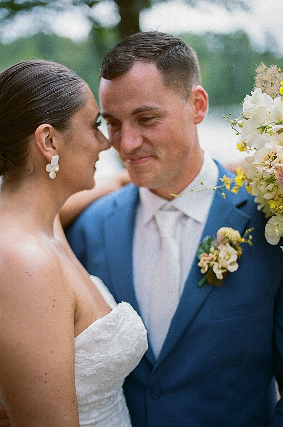 Wedding couple portrait close up bride and groom embracing with forehead touch, strapless lace dress and blue suit amid soft greenery trees