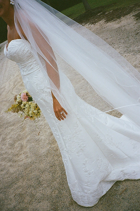 Wedding dress close-up showing strapless lace wedding dress, long veil and bouquet, with a wedding ring detail on a sandy shoreline
