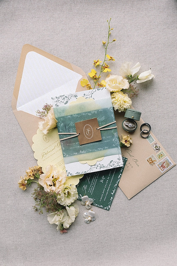 Invitation suite wedding invitation flat lay with vellum wrap, wax seal, twine, stamps, rings, and pearl earrings on linen backdrop