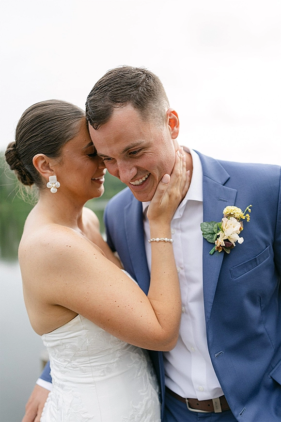 Couple portrait of bride and groom close up as she touches his face, her pearl statement earrings shining by a lakeside backdrop