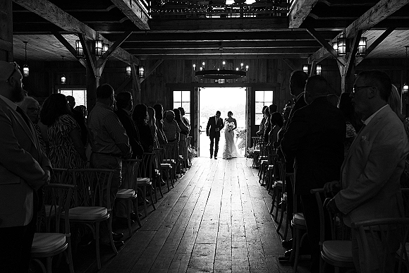 Wedding recessional as bride and groom walk down the barn aisle, guests lining chairs; chandelier overhead, backlit doorway ahead
