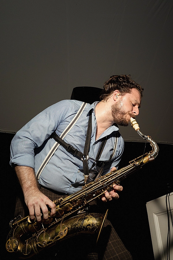 Wedding saxophonist, wedding live musician in suspenders and button-down, playing saxophone on an indoor reception stage against a dark wall