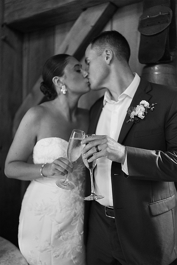 Wedding kiss portrait of newlyweds kissing while holding champagne flutes, bride in strapless lace dress and groom in suit by wooden beams