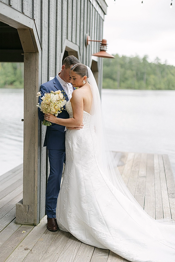 Couple portrait of bride and groom embrace, her long veil and bouquet beside a wooden boathouse on a dock by the lake