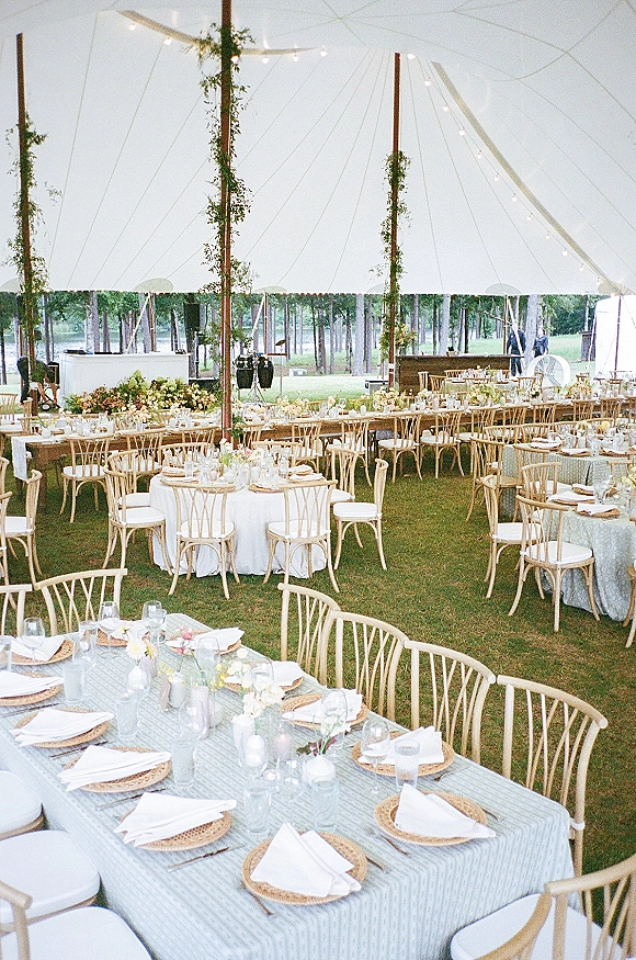 Wedding reception decor under a sailcloth tent reception with string lights, long banquet and round tables set with candles and florals on a lawn