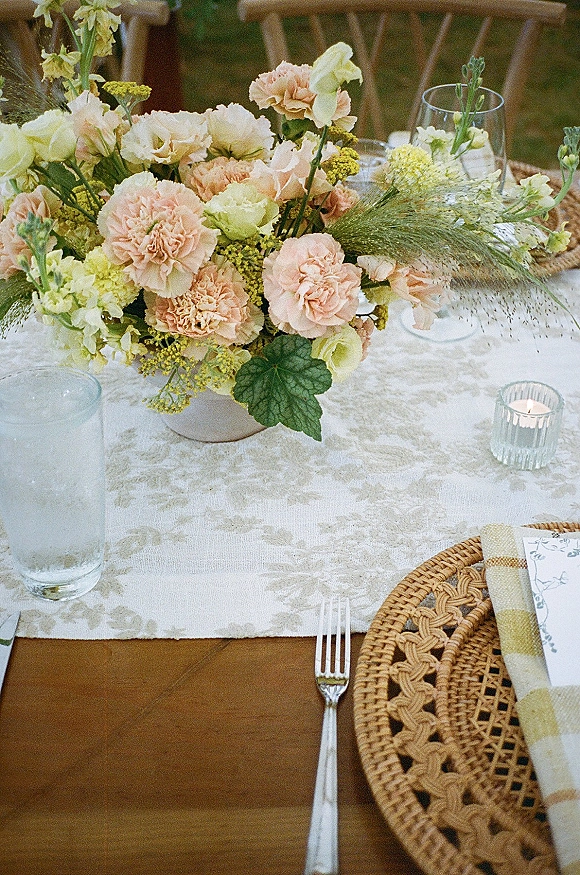 Reception tablescape with wedding table centerpiece of carnations and baby’s breath on a wooden table with rattan charger, glasses, and candle on lawn