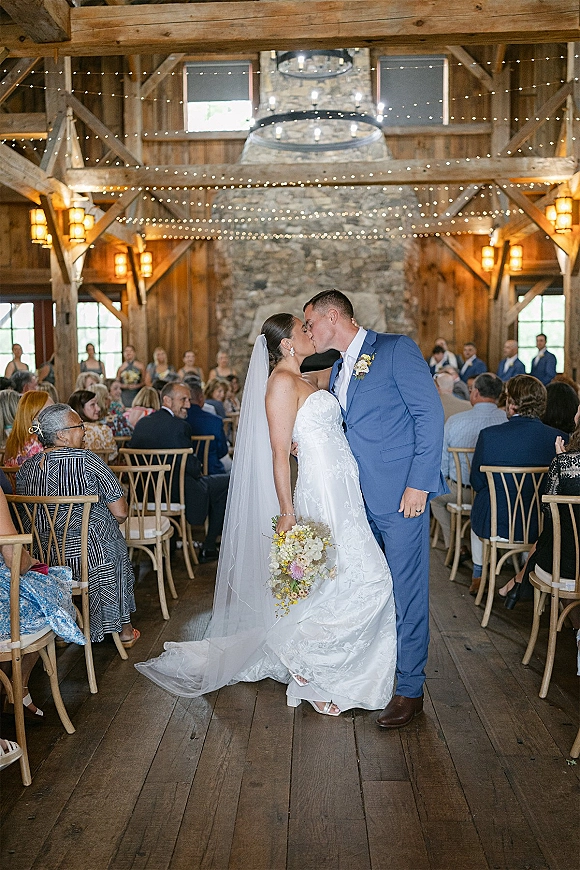 Wedding kiss as bride in lace dress and veil holds bouquet, groom in blue suit, in barn aisle with guests, string lights and fireplace