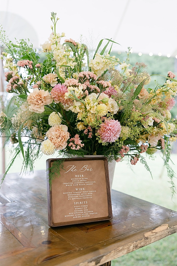 Wedding bar sign with a framed bar menu beside a lush floral arrangement on a wooden table under a white tent canopy with string lights