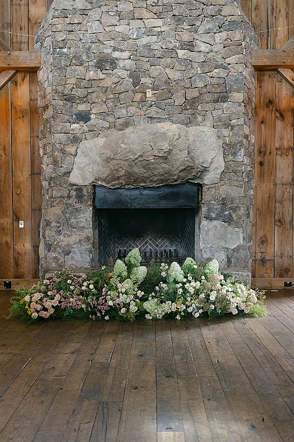 Fireplace floral arrangement with white and blush blooms and greenery along a stone hearth, set against wood-paneled walls and floor