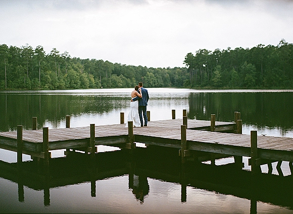 Couple portrait of newlyweds on dock, embracing from behind with wedding dress and groom suit by a reflective lake and forest backdrop