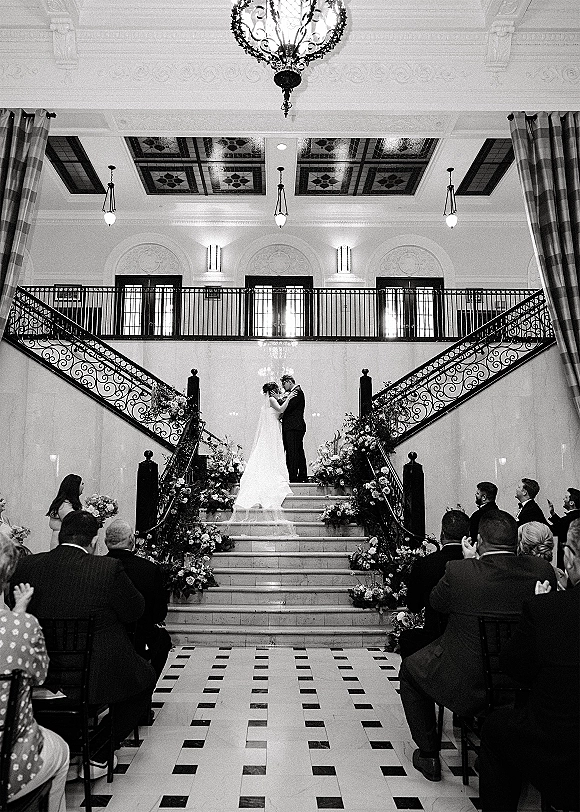 Wedding ceremony kiss as bride and groom embrace on a grand staircase, chandelier overhead, guests seated beside floral arrangements and veil