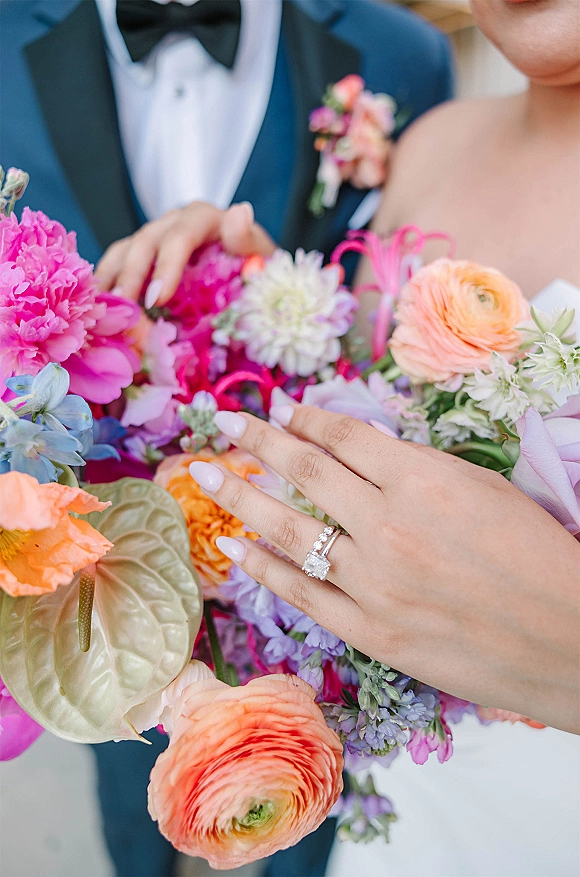 Engagement ring close-up with wedding ring stack on manicured hand holding a bright pink bouquet, blurred couple against neutral backdrop