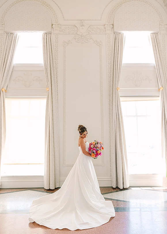 Bridal portrait of a bride holding bouquet in a strapless gown with long train, looking down by arched windows and tall drapery in soft light