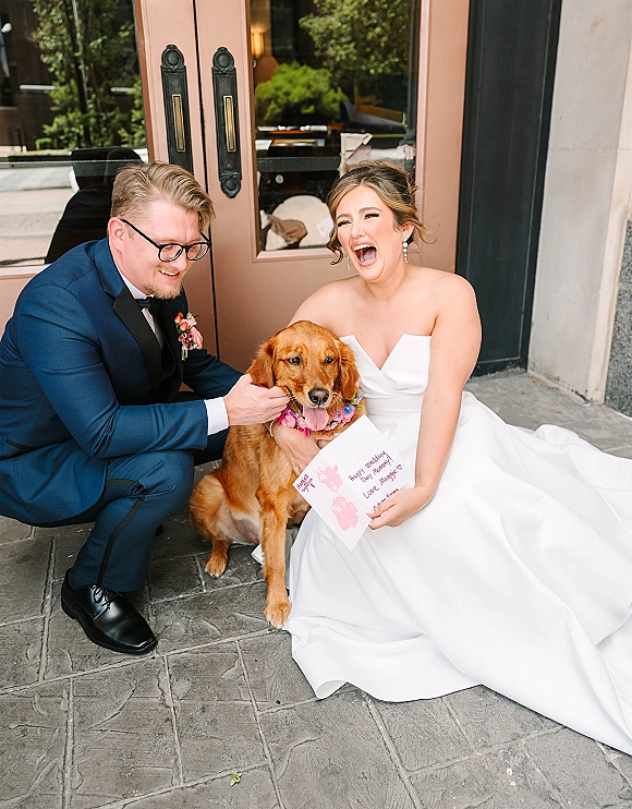 Wedding couple with dog posing with a golden retriever in a floral collar, bride in strapless gown and groom in navy tux by glass doors