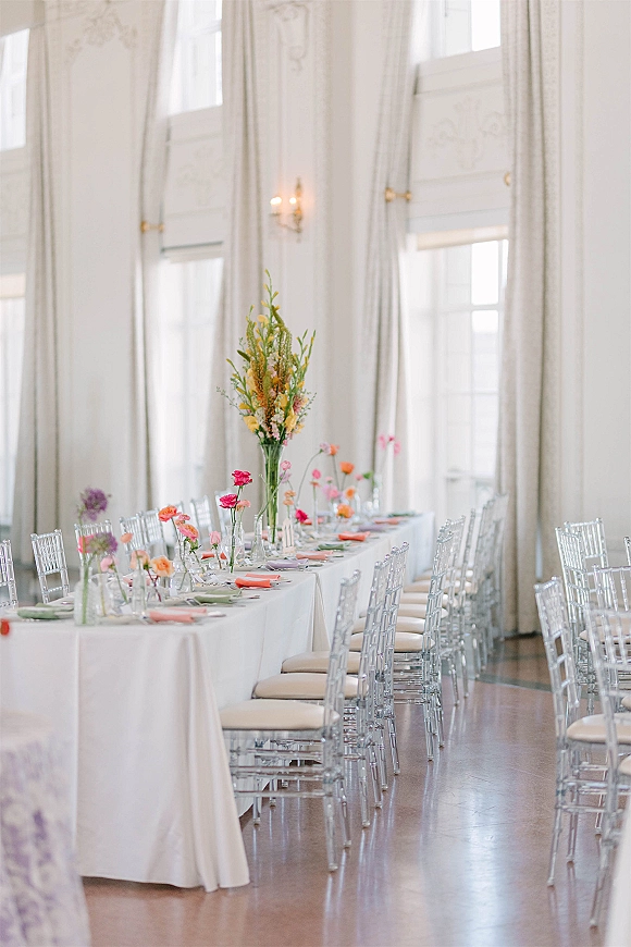 Reception tablescape with a long banquet table, white tablecloth, bud vases and tall colorful centerpiece, coral napkins by tall windows