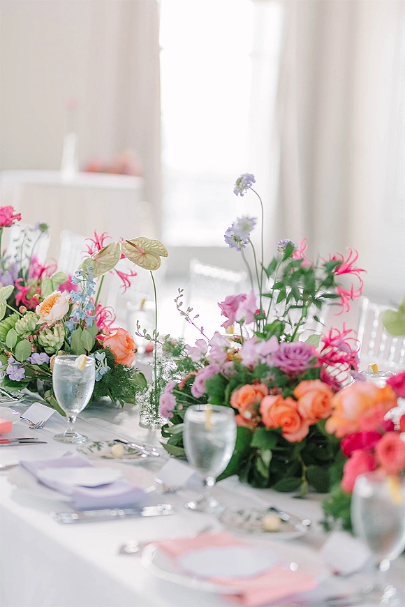 Reception tablescape with wedding table centerpiece of colorful florals and greenery garland, lavender and coral napkins by bright windows