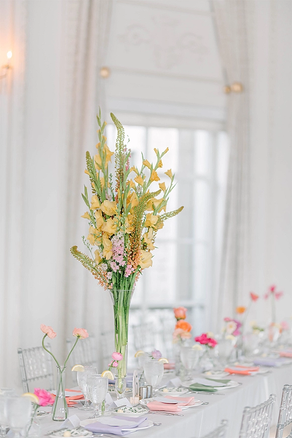 Reception tablescape with tall floral centerpiece, bud vases, pastel napkins and place cards, set by candlelight near large windows and sheer drapes