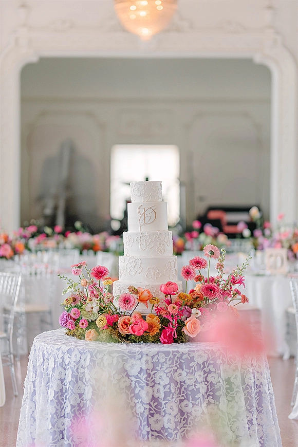 Wedding cake table with a tiered white wedding cake, lace detailing and monogram topper, surrounded by vibrant florals under a chandelier in a reception hall