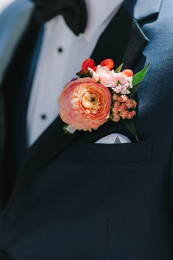 Groom boutonniere with peach ranunculus pinned to a black tux lapel, paired with a bow tie and white pocket square close-up