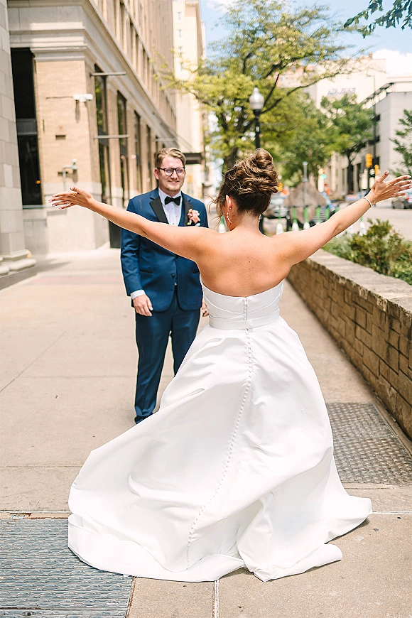 Wedding first look as the bride reveals dress with arms open, approaching a groom in tuxedo and glasses on a city sidewalk by stone building