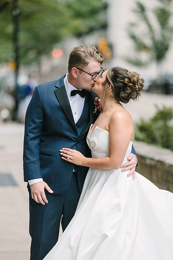 Wedding kiss portrait of bride and groom kissing on a city sidewalk, bride in strapless gown, groom in navy tuxedo with glasses and boutonniere
