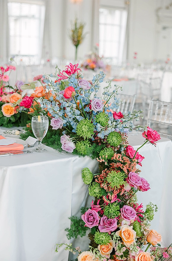 Wedding reception centerpiece with head table floral runner of roses and delphinium on white linens in a bright windowed room