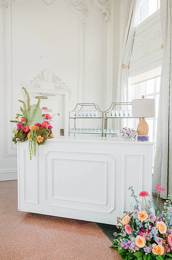 Wedding bar setup with white bar, coral-pink floral accents, drink menu sign, and glassware under tall windows and ornate molding