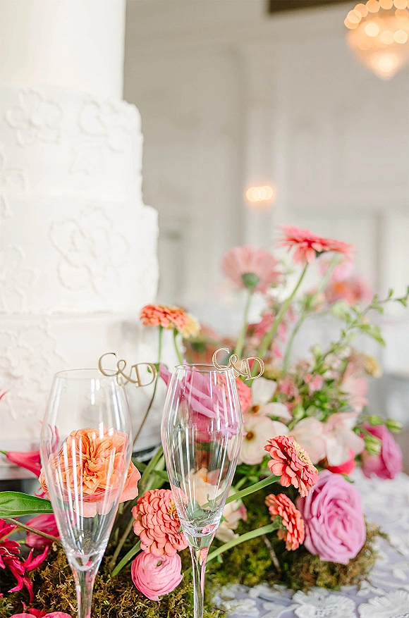 Wedding cake with white textured frosting, tiered design and initials topper, flanked by champagne flutes and pink-coral flowers on a table indoors