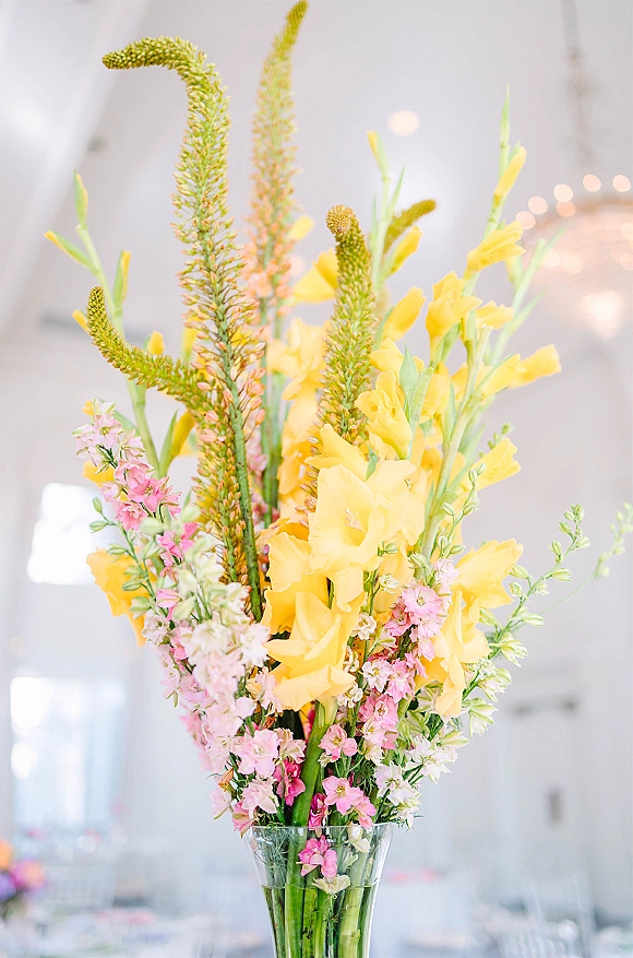 Wedding floral centerpiece in a tall vase with yellow and pink blooms and greenery stems under bistro lights in a white tent reception