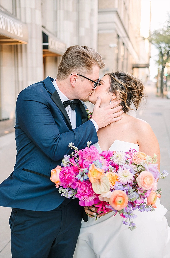 Wedding kiss portrait of bride and groom kissing, groom hand on her face, navy tuxedo and strapless dress on a city sidewalk backdrop