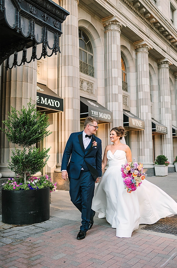 Couple portrait of bride and groom walking hand in hand on a city sidewalk, bride in veil with bright pink bouquet beside columns