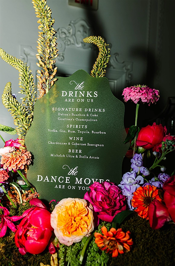 Wedding bar sign with a signature drinks sign in painted lettering, framed by peonies, zinnias, greenery, and moss on a molded wall