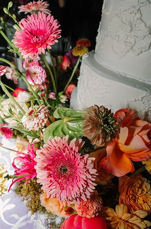 Wedding cake with white buttercream tiers and piped floral icing, styled with gerbera daisies, roses, zinnias, and moss on a white tablecloth