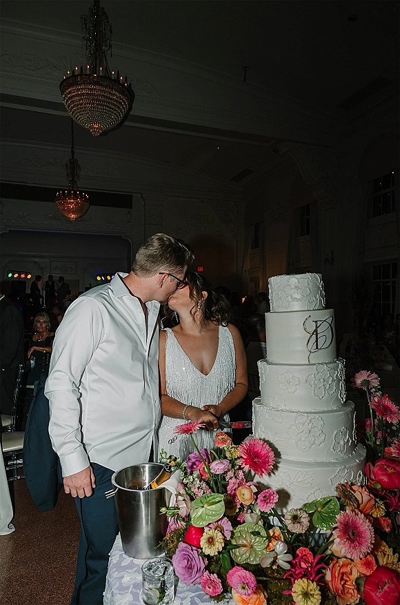 Wedding cake cutting as the bride and groom kiss beside a four-tier cake with bright florals and a champagne bucket under chandeliers