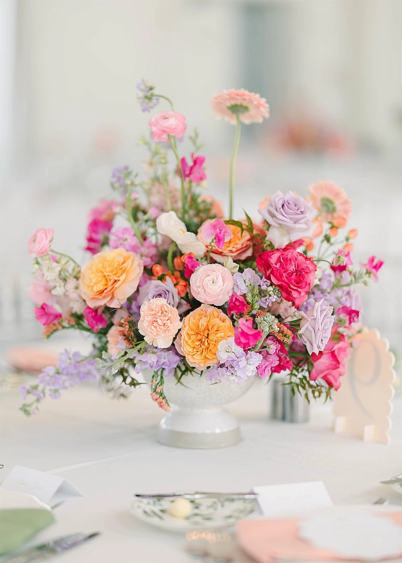 Wedding centerpiece with colorful wedding centerpiece blooms in a ceramic compote vase, set beside place cards and silverware on a reception table