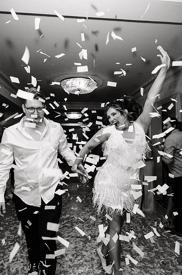 Wedding exit as bride in fringe mini dress and groom hold hands under confetti in chandelier-lit hallway with cheering guests