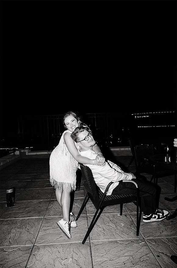 Couple portrait in a black and white wedding photo, bride hugging groom on an outdoor patio at night with city lights behind them