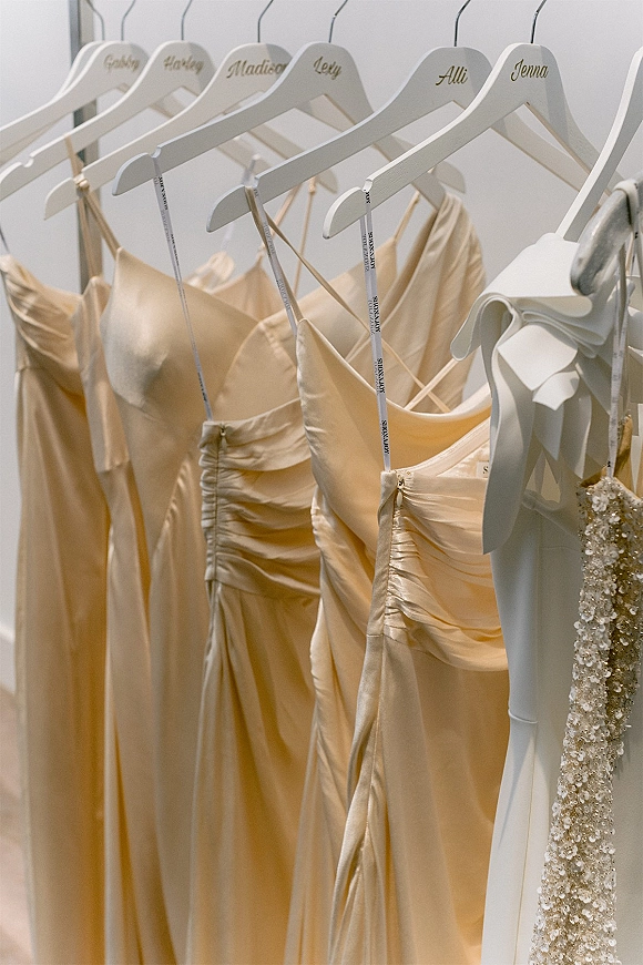 Bridesmaid dresses in champagne tones hanging on personalized white wooden hangers on a closet rack against a white wall