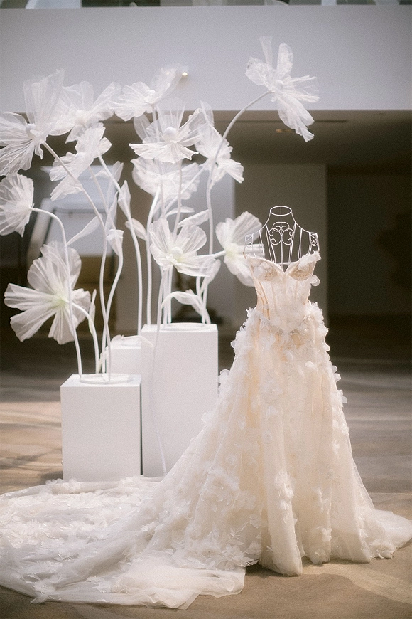 Wedding dress displayed on a mannequin, strapless lace gown with floral appliqué and long train, framed by white floral pedestals indoors