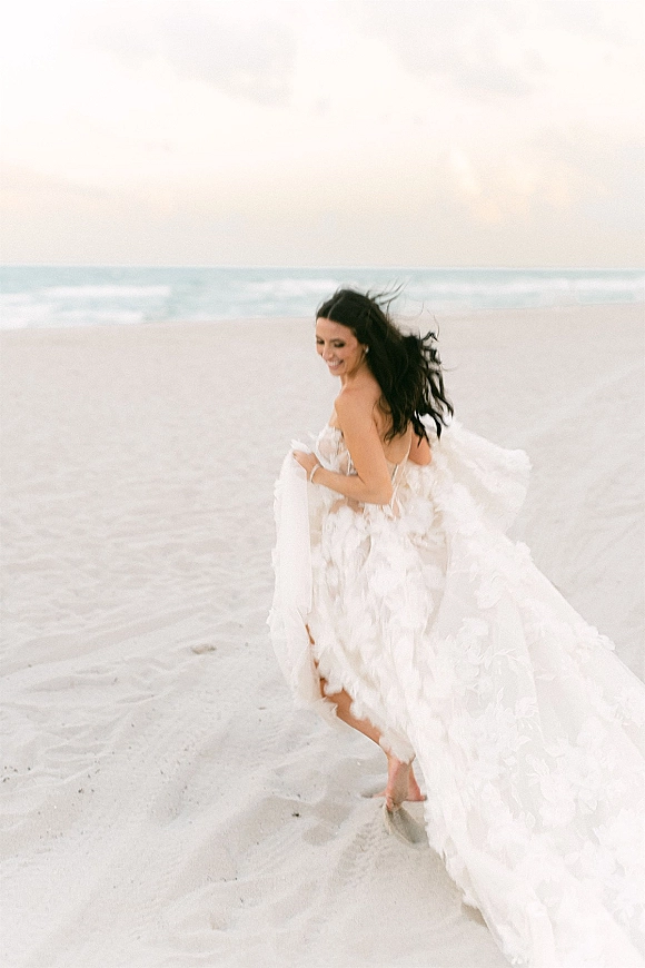 Bridal portrait of a bride running barefoot on the beach in a strapless floral appliqué gown with flowing train, laughing by the ocean horizon
