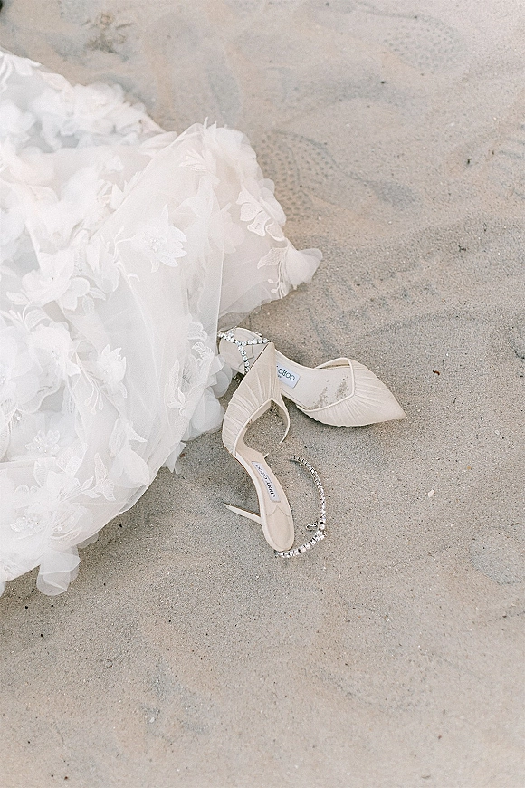 Bridal shoes with crystal ankle straps and pointed toes resting on beach sand beside lace appliqué and tulle from the gown train
