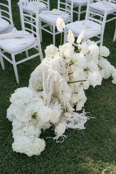 Ceremony aisle flowers in a ground floral arrangement of white roses, hydrangeas and anthurium beside Chiavari chairs with paper fans on a grass lawn