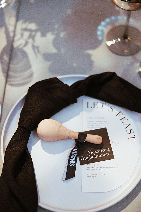 Wedding place setting with black napkin place setting on a white plate, round menu card, place card, and wooden maraca favor on white tablecloth