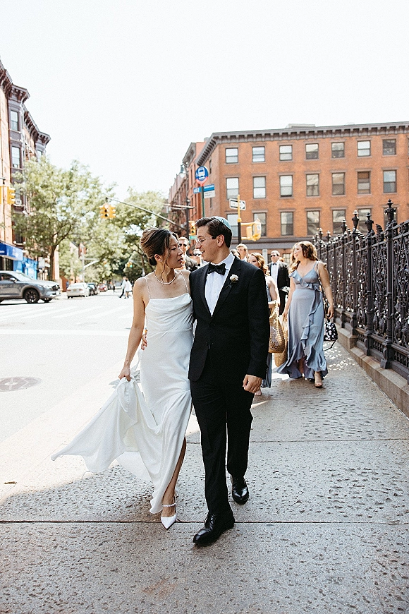 Couple portrait of bride and groom walking on a city street, bride lifting her dress train as bridesmaids follow behind