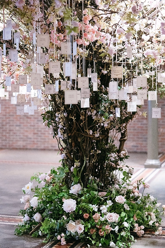Wedding seating chart with hanging escort cards on ribbons, framed by flowering branches and blush-and-white roses against a brick wall backdrop
